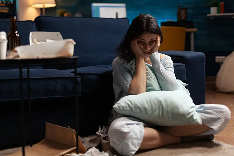 young woman sitting on the floor with her hands on her face, empty to-go food boxes on the nearby table